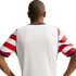 Rear view of a man with a buzz cut wearing a white jersey with red-and-white patterned sleeves and navy trim, standing with hands on hips against a black background.