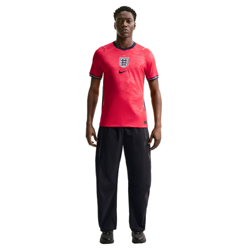 A man stands in a red England national football team jersey and black trousers against a plain black background.