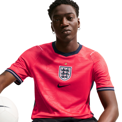 A man wearing a red England national football team jersey with the Three Lions crest and Nike logo, holding a white football against a black background.