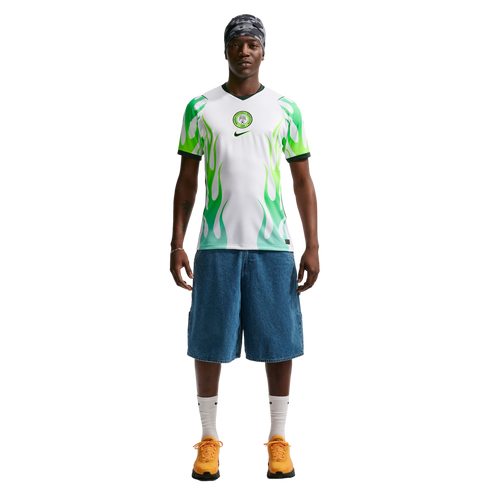 A man wearing a white and green flame-patterned soccer jersey, denim shorts, and orange sneakers stands against a black background.