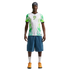 A man wearing a white and green flame-patterned soccer jersey, denim shorts, and orange sneakers stands against a black background.