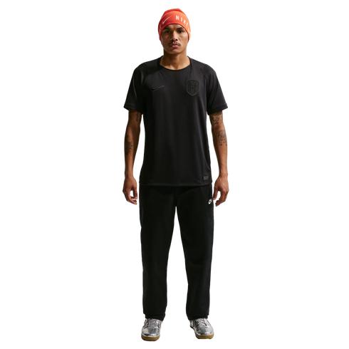 A man wearing an orange Nike beanie, a black t-shirt and sweatpants, and silver sneakers stands against a black background.