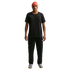 A man wearing an orange Nike beanie, a black t-shirt and sweatpants, and silver sneakers stands against a black background.