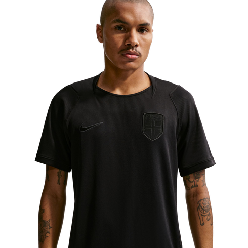 A man with a buzz cut and forearm tattoos wears a black Nike athletic shirt with a subtle monochromatic shield crest, standing against a dark background.