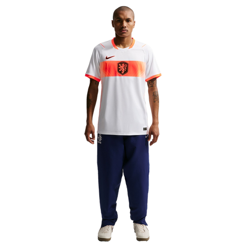 A full-length studio portrait of a man standing against a black background. He is wearing a white Nike soccer jersey with an orange horizontal band across the chest that features a black lion crest. He is also wearing navy blue tracksuit pants and off-whi