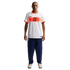 A full-length studio portrait of a man standing against a black background. He is wearing a white Nike soccer jersey with an orange horizontal band across the chest that features a black lion crest. He is also wearing navy blue tracksuit pants and off-whi