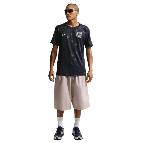 A man models a dark blue Nike USA soccer jersey with grey stars, oversized light pink shorts, and blue sneakers against a black background.