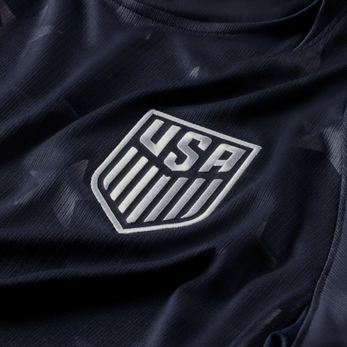Close-up of a navy blue USA soccer jersey featuring a silver embroidered national team crest and subtle star patterns on the fabric.