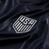 Close-up of a navy blue USA soccer jersey featuring a silver embroidered national team crest and subtle star patterns on the fabric.