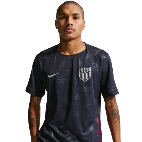 A man wearing a dark blue Nike USA soccer jersey with a subtle star pattern and red trim. He has tattoos on his arms and stands against a black background.