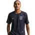 A man wearing a dark blue Nike USA soccer jersey with a subtle star pattern and red trim. He has tattoos on his arms and stands against a black background.
