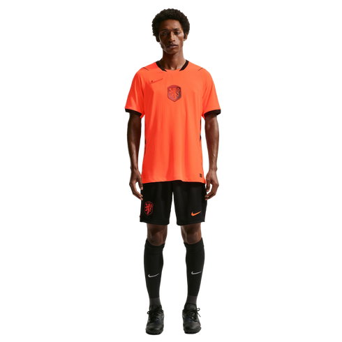 A male model stands against a black background wearing a bright orange Netherlands national team soccer jersey with black trim, matching black shorts, high black socks, and black cleats.