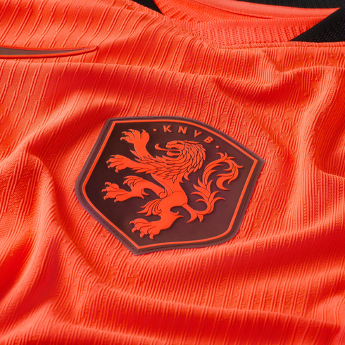 A close-up of a bright orange textured soccer jersey featuring the KNVB crest. The dark, shield-shaped patch displays an orange lion in a rampant pose with the letters "K N V B" above it. The jersey's fabric has a distinctive vertical ribbed pattern.