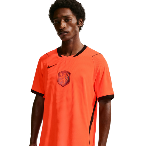 A young man with dark skin and curly hair wears a bright orange Netherlands national team home jersey. The jersey has a black V-neck collar, sleeve trim, and a black Nike swoosh on the chest, with the KNVB lion crest in the center. He is positioned agains