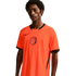 A young man with dark skin and curly hair wears a bright orange Netherlands national team home jersey. The jersey has a black V-neck collar, sleeve trim, and a black Nike swoosh on the chest, with the KNVB lion crest in the center. He is positioned agains