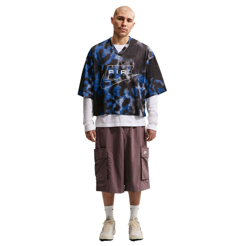 A man wears an oversized blue and black patterned Nike jersey, white long-sleeve shirt, baggy brown cargo shorts, and cream sneakers against a white background.