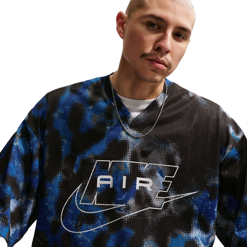 A person with a shaved head wears a blue and black patterned mesh Nike "AIR" jersey and a silver chain against a white background.
