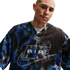 A person with a shaved head wears a blue and black patterned mesh Nike "AIR" jersey and a silver chain against a white background.