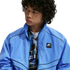 Young man with dark curly hair wearing a bright blue Nike Air windbreaker jacket over a patterned shirt, looking to the side against a white background.
