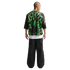 Back view of a person wearing a green and black mesh top over a white tee and baggy black trousers against a white backdrop.