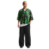 A man with a shaved head wearing a patterned green and black Nike mesh jersey, baggy black pants, and black sneakers against a white background.