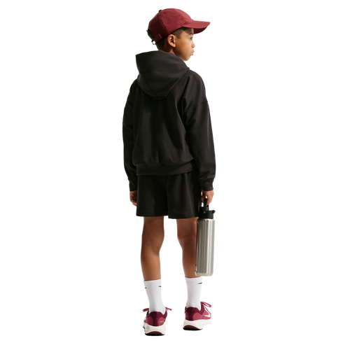 Back view of a child in a maroon cap, black hoodie, and shorts. They hold a silver water bottle and wear maroon sneakers against a black background.