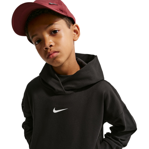 A boy wearing a black Nike hoodie and a maroon Nike baseball cap looks at the camera against a black background.