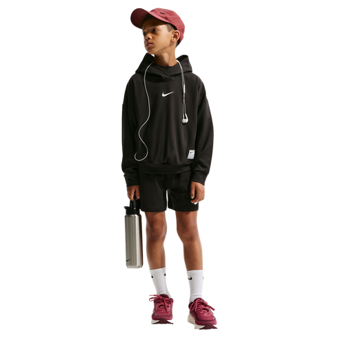 A young boy in a black Nike hoodie, black shorts, maroon cap, and maroon sneakers stands against a black background, holding a silver water bottle and wearing wired earphones.