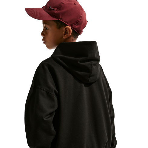 Side view of a young boy looking over his shoulder, wearing a maroon baseball cap and a black hoodie against a plain black background.