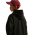 Side view of a young boy looking over his shoulder, wearing a maroon baseball cap and a black hoodie against a plain black background.