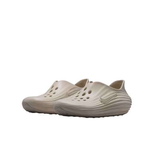 A pair of tan Nike foam clogs featuring perforated holes and wavy textured sides, displayed against a solid black background.