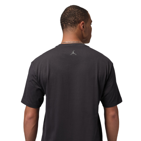 Rear view of a man wearing a charcoal gray Jordan t-shirt with a small silver Jumpman logo on the upper back. He is set against a solid black background.
