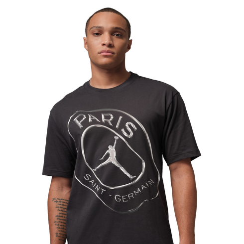 A man wearing a black t-shirt with a metallic silver graphic of the Paris Saint-Germain logo and Jordan Brand Jumpman on a black background.