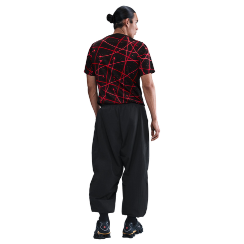 Back view of a man wearing a black t-shirt with a red abstract pattern and baggy black pants against a white background.