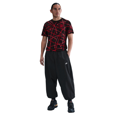 A man in a red and black patterned soccer jersey, baggy black pants, and sneakers, standing against a white background.
