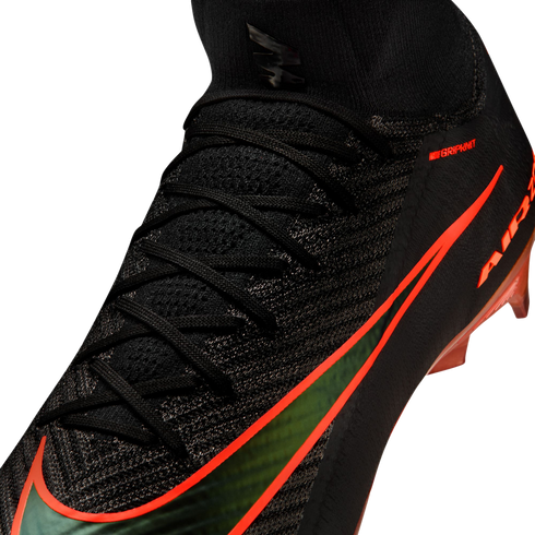 A close-up view of a black Nike soccer cleat with a textured knit upper and a sock-like collar. The shoe features a large, iridescent green Nike swoosh outlined in bright orange, along with small orange "NIKE GRIPKNIT" text and orange studs visible at the