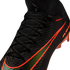 A close-up view of a black Nike soccer cleat with a textured knit upper and a sock-like collar. The shoe features a large, iridescent green Nike swoosh outlined in bright orange, along with small orange "NIKE GRIPKNIT" text and orange studs visible at the