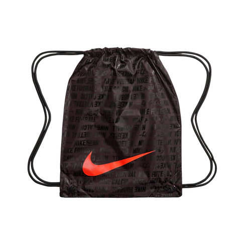 A dark brown Nike drawstring bag featuring a large, bright red swoosh across the center. The bag is covered in a repeating black text pattern of "NIKE" followed by translations for the word "football" in various languages and scripts. Black cord drawstrin