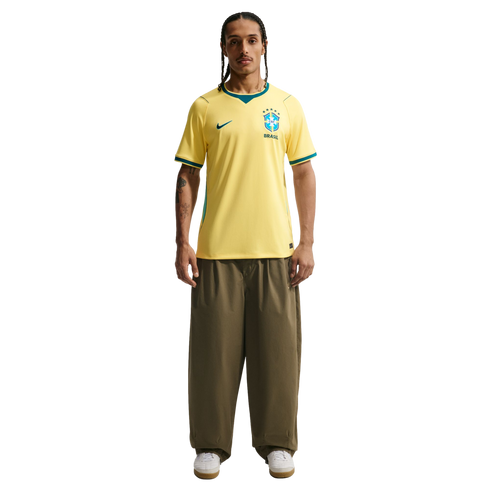 A man stands against a black background wearing a yellow Nike Brazil national team soccer jersey, olive green wide-leg trousers, and white sneakers. He has braided hair, a mustache, and tattoos on his right arm.
