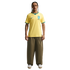 A man stands against a black background wearing a yellow Nike Brazil national team soccer jersey, olive green wide-leg trousers, and white sneakers. He has braided hair, a mustache, and tattoos on his right arm.