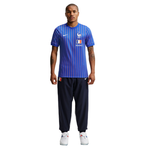A man wearing a blue pinstriped Nike x Jacquemus France soccer jersey, navy track pants, and white sneakers stands against a plain white background.