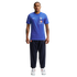 A man wearing a blue pinstriped Nike x Jacquemus France soccer jersey, navy track pants, and white sneakers stands against a plain white background.