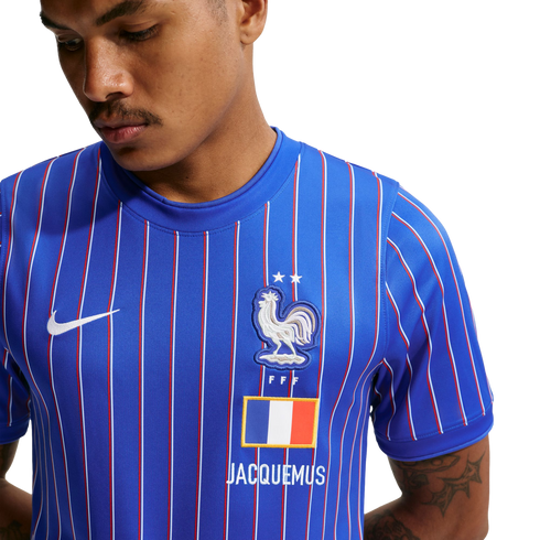 Close-up of a man wearing a blue Nike x Jacquemus France football jersey with red and white pinstripes, featuring the FFF rooster crest, two stars, and a French flag.