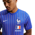 Close-up of a man wearing a blue Nike x Jacquemus France football jersey with red and white pinstripes, featuring the FFF rooster crest, two stars, and a French flag.
