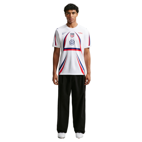 A man stands wearing a white USA soccer jersey with red and blue accents, black wide-leg trousers, and white sneakers against a plain white background.