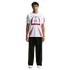 A man stands wearing a white USA soccer jersey with red and blue accents, black wide-leg trousers, and white sneakers against a plain white background.