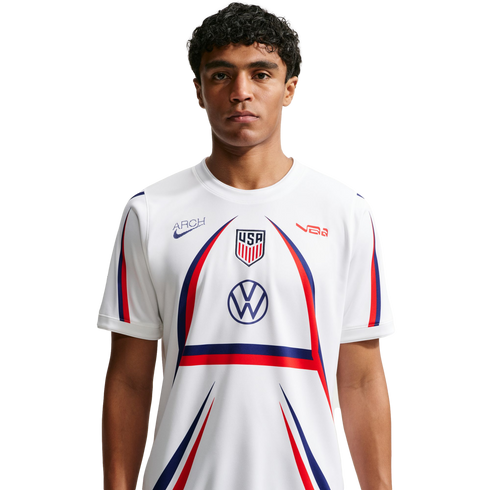 A man with curly hair wears a white USA soccer jersey featuring red and blue accents and a large arch design, centered against a plain white background.