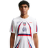 A man with curly hair wears a white USA soccer jersey featuring red and blue accents and a large arch design, centered against a plain white background.