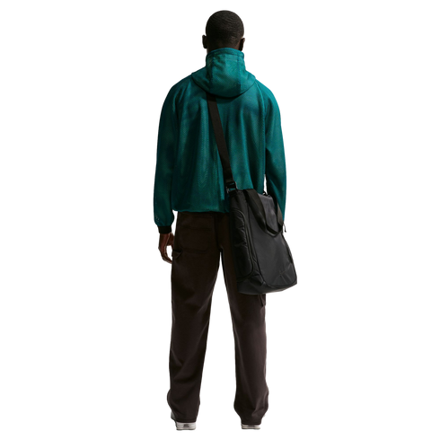 Rear view of a man standing against a solid black background, wearing a teal green mesh hooded jacket, dark brown trousers, and white sneakers. A large black messenger bag is slung over his right shoulder.
