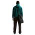 Rear view of a man standing against a solid black background, wearing a teal green mesh hooded jacket, dark brown trousers, and white sneakers. A large black messenger bag is slung over his right shoulder.